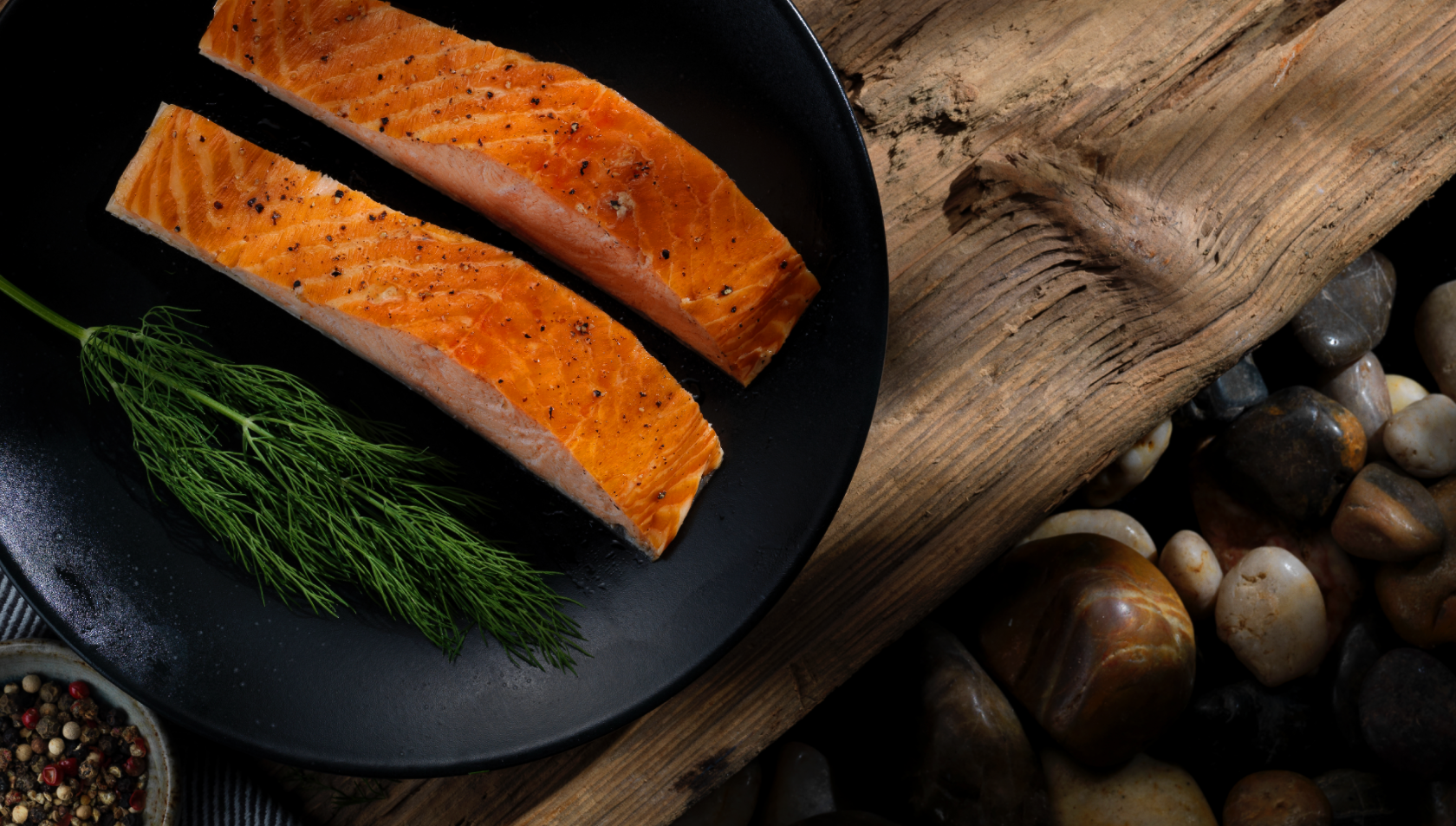 Two fillets of salmon with cracked black pepper and some fresh dill, beautifully presented on a black dinner plate.